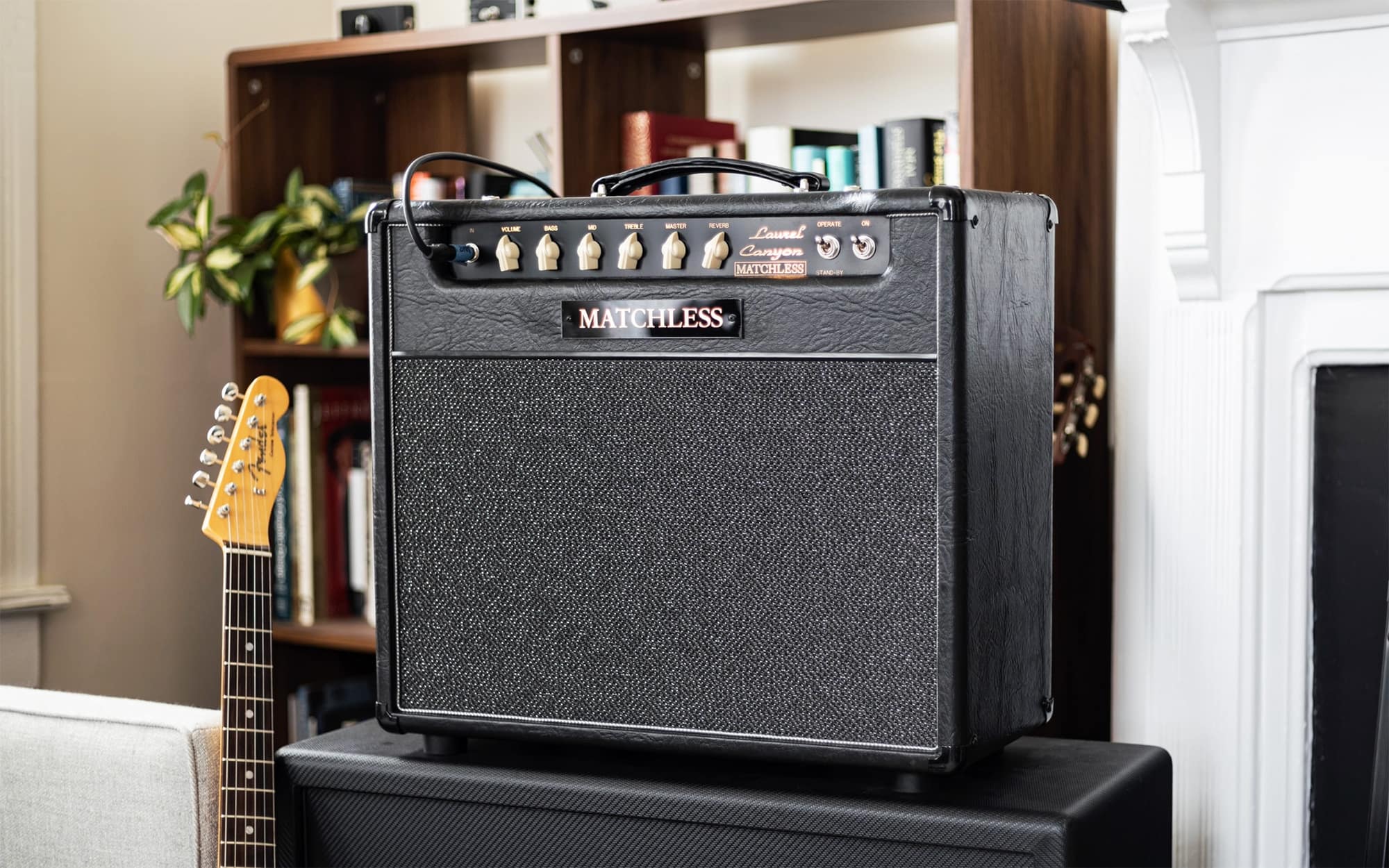 A black Matchless guitar amplifier with a textured grille and control knobs, sitting on top of a speaker in a home studio or living room setting with a guitar visible in the background.